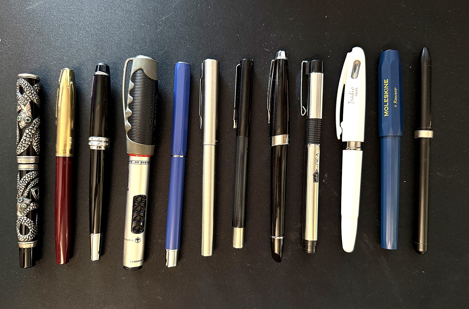 Twelve fountain pens laid side by side on a dark desk mat, including the silver Parker Snake, a burgundy and gold Sheaffer, a Rotring, a Pentel Tradio and a blue Moleskine x Kaweco.